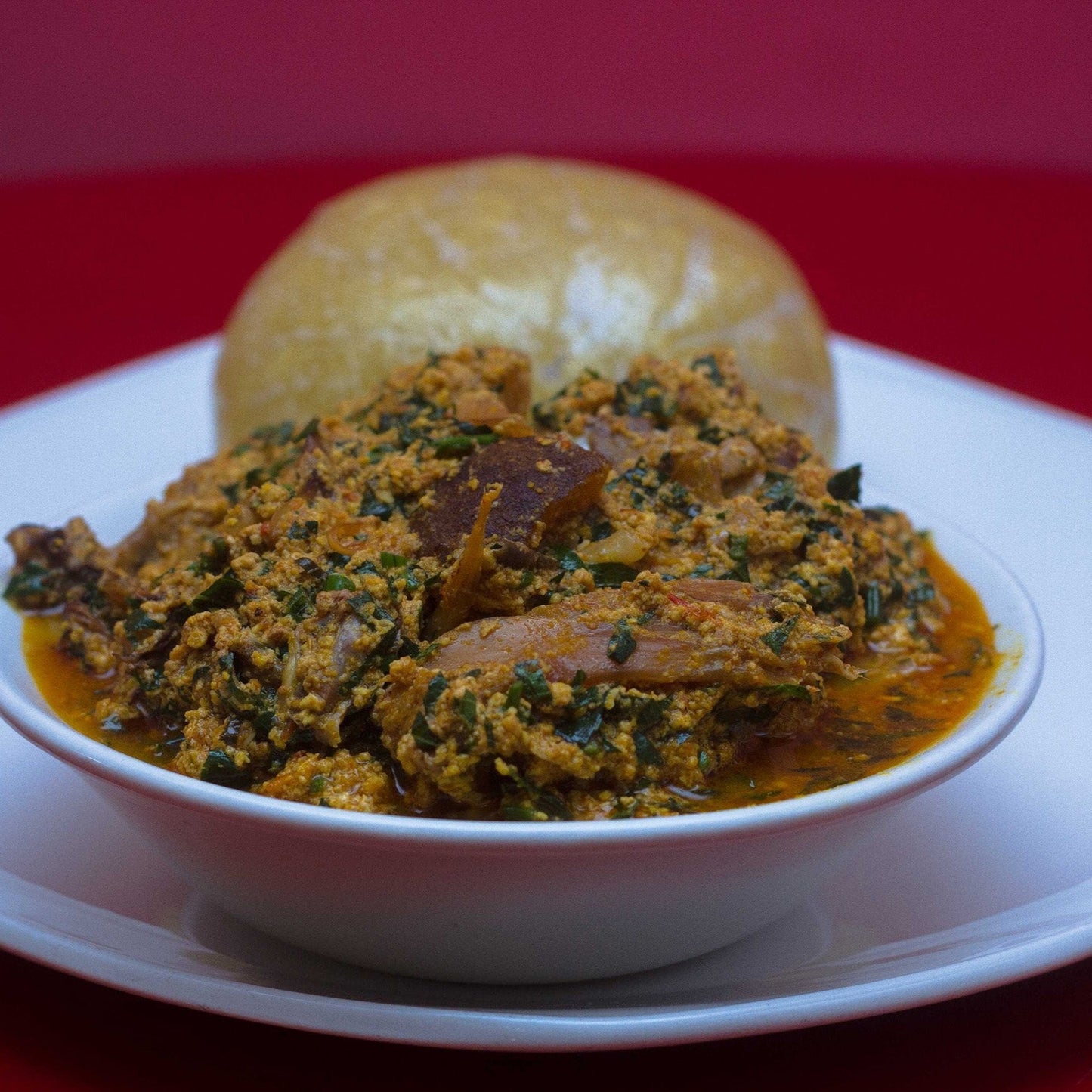 A bowl of Egusi Soup, with visible chunks of meat and leafy greens, served on a white plate against a red background.